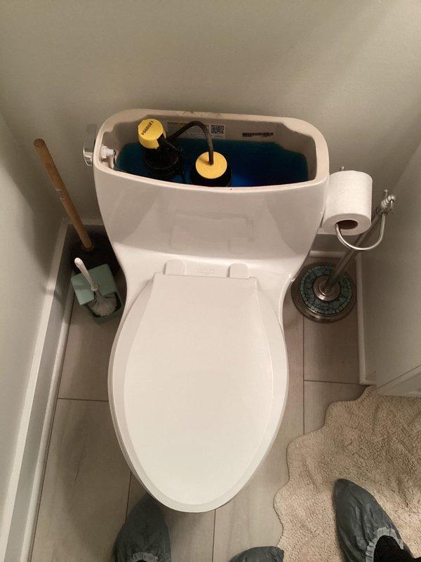 Responded to complaint of toilet tank not fully draining on first flush, an issue present since installation several years ago in 65-year-old home. Upon inspection, observed dual-flush toilet mechanism with blue-tinted water in tank and two yellow float devices connected by tubing. Examined internal flush valve, fill valve, and flapper assembly for proper operation and adjustment. Photographed tank components and related plumbing system including water heater installation to document existing conditions. Advised customer on findings and available options for resolving incomplete flush cycle.