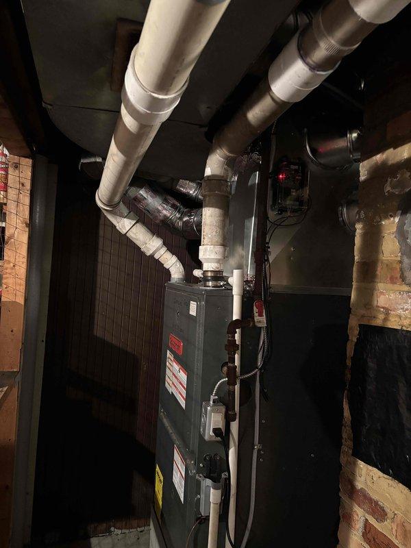 Complete air conditioning system replacement performed at residential property. Removed existing HVAC equipment and installed new outdoor condenser unit on composite pad against exterior wall with proper clearances. Unit features cylindrical top section with protective mesh grilling and horizontal louvered side panels. Mounted electrical disconnect box on siding and connected gas line with appropriate regulator and black piping. Installed white PVC condensate drain lines with proper routing and elbow fittings along exterior wall.

Inside mechanical room, installed new air handler unit with proper ductwork connections to existing distribution system. Connected multiple branch ducts including insulated and metallic ductwork extending from top of unit. Verified all manufacturer specifications, warning labels, and informational plates properly affixed to equipment. Completed all electrical connections, refrigerant line sets, and condensate drainage. System tested and verified operational w