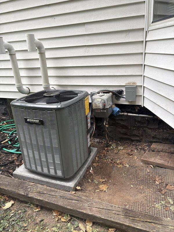 Complete air conditioning system replacement performed at residential property. Removed existing HVAC equipment and installed new outdoor condenser unit on composite pad against exterior wall with proper clearances. Unit features cylindrical top section with protective mesh grilling and horizontal louvered side panels. Mounted electrical disconnect box on siding and connected gas line with appropriate regulator and black piping. Installed white PVC condensate drain lines with proper routing and elbow fittings along exterior wall.

Inside mechanical room, installed new air handler unit with proper ductwork connections to existing distribution system. Connected multiple branch ducts including insulated and metallic ductwork extending from top of unit. Verified all manufacturer specifications, warning labels, and informational plates properly affixed to equipment. Completed all electrical connections, refrigerant line sets, and condensate drainage. System tested and verified operational w