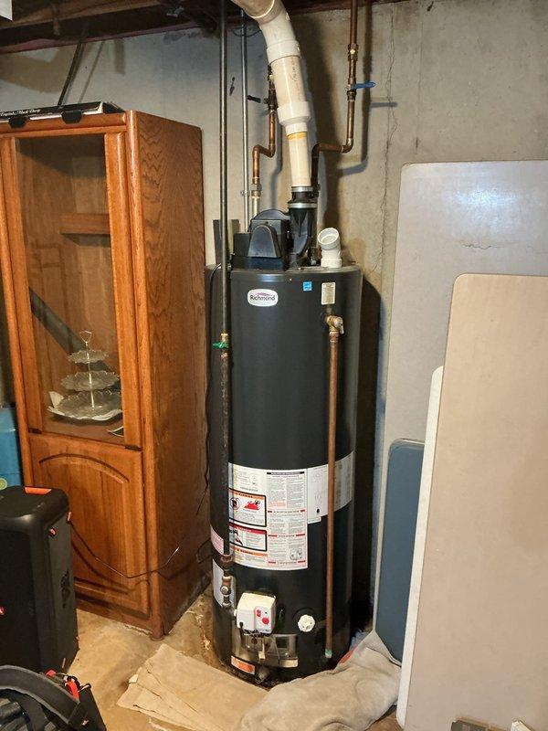 Completed estimate for water heater replacement in basement utility area. Assessed existing black cylindrical water heater with copper supply lines, PVC venting system, and associated shut-off valves mounted against concrete block wall. Documented current plumbing configuration including hot and cold water connections with brass fittings, blue and black-handled isolation valves, white PVC drain line, and rubber coupling connections at base. Photographed installation layout and clearance requirements for equipment replacement recommendation.