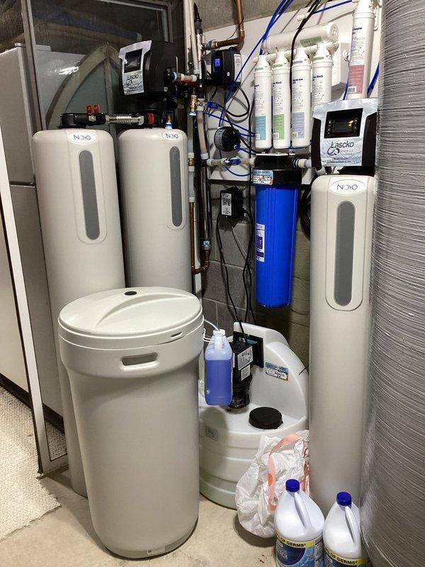 Responded to service call for inspection of complex residential water treatment system. Upon arrival, evaluated multi-component setup including multiple water softener tanks, whole-house filtration system with wall-mounted filter cartridge assembly, pressure storage tank, and interconnected plumbing infrastructure. Documented system configuration with photographs showing N2O-labeled softener units, blue filter housing, copper piping network, pressure gauges, control boxes, and chemical treatment bottles. Assessed system complexity and serviceability to determine if equipment falls within company's scope of maintenance capabilities.
