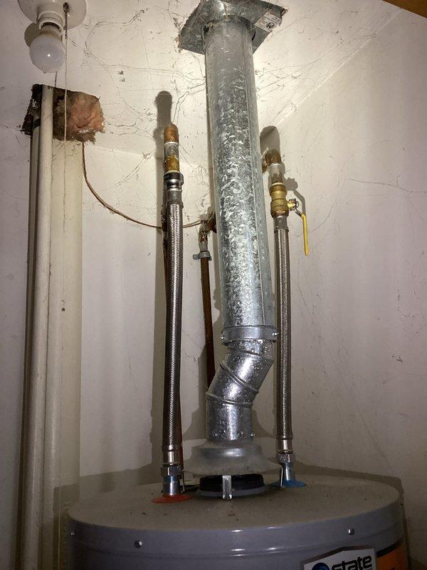 Provided estimate for replacement of existing Ostate brand water heater, approximately 20 years old. Unit is installed in confined utility closet with vertical exhaust venting through ceiling. Existing installation shows significant age with deteriorated vent insulation, cracked wall surfaces, and worn components throughout. Documented current configuration including flexible water supply lines with shut-off valves and existing venting system for replacement specifications.