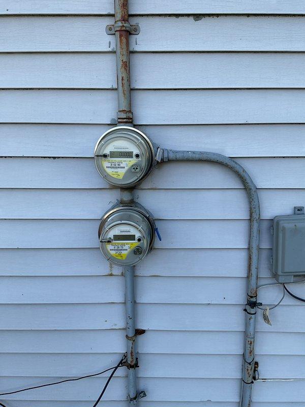 Conducted electrical estimate for century-old home experiencing hazardous conditions with crackling and warm fuse box affecting entire electrical system. Documented exterior electrical service equipment including dual utility meters, weathered conduit system with visible rust and corrosion at connection points, and aging infrastructure requiring comprehensive evaluation for safe electrical panel replacement and service upgrade.