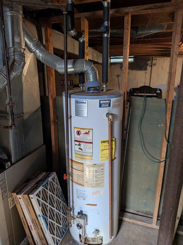 Provided estimate for water heater replacement in residential basement utility area. Assessed existing white cylindrical water heater with connected flue piping and adjacent HVAC ductwork system. Documented current equipment specifications and installation configuration for replacement proposal.