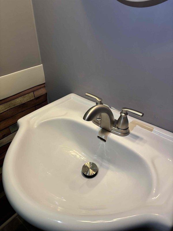 Completed comprehensive plumbing repairs in residential bathroom and basement utility area. Serviced bathroom sink faucet assembly with brushed nickel finish and dual lever handles, ensuring proper operation and addressing water flow issues. Inspected and cleared drain stopper mechanism in porcelain basin. In basement area, addressed corroded valve assembly at base of vertical PVC drainage line along foundation wall. Replaced heavily oxidized green valve fitting showing moisture damage and age-related deterioration. Verified proper drainage function and checked for leaks throughout system after repairs were completed.