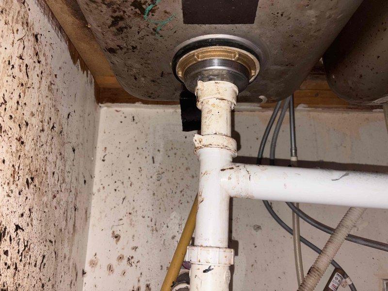 Responded to plumbing leak issue involving broken sink drain piping. Upon inspection, found deteriorated PVC drain assembly beneath sink fixture with compromised connections showing signs of age, discoloration, and potential failure points at joints. Assessed white PVC pipe configuration extending from sink drain through floor penetration into crawl space below. Documented existing conditions including corroded drain gasket, stained pipe connections, and evidence of moisture intrusion on surrounding surfaces. Evaluated structural mounting points and confirmed proper vertical alignment of drain stack through floor assembly with chrome flange securing pipe penetration. Addressed leak source at drain connection points and replaced failed components to restore proper drainage function and eliminate water discharge into cabinet and crawlspace areas.