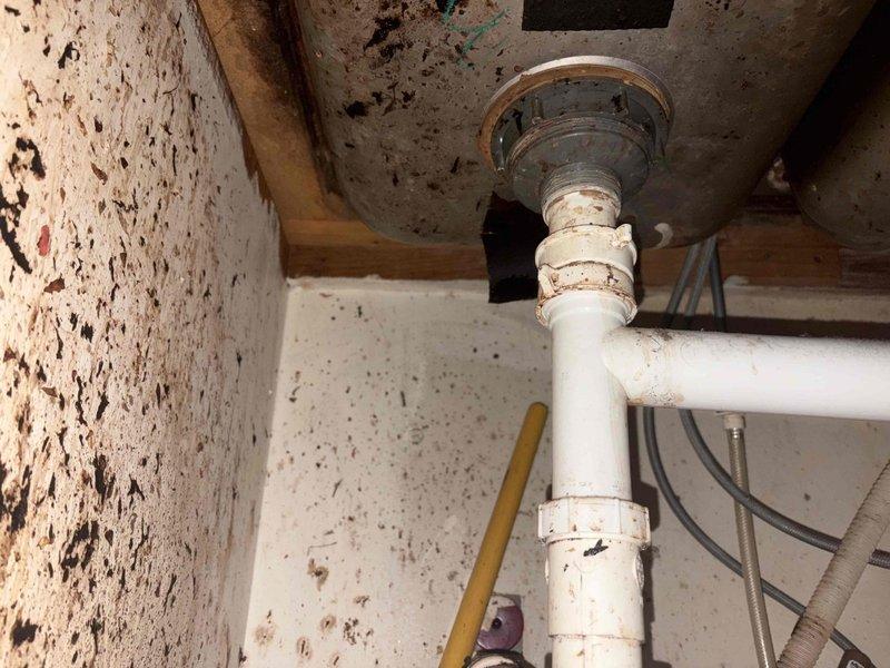 Responded to plumbing leak issue involving broken sink drain piping. Upon inspection, found deteriorated PVC drain assembly beneath sink fixture with compromised connections showing signs of age, discoloration, and potential failure points at joints. Assessed white PVC pipe configuration extending from sink drain through floor penetration into crawl space below. Documented existing conditions including corroded drain gasket, stained pipe connections, and evidence of moisture intrusion on surrounding surfaces. Evaluated structural mounting points and confirmed proper vertical alignment of drain stack through floor assembly with chrome flange securing pipe penetration. Addressed leak source at drain connection points and replaced failed components to restore proper drainage function and eliminate water discharge into cabinet and crawlspace areas.