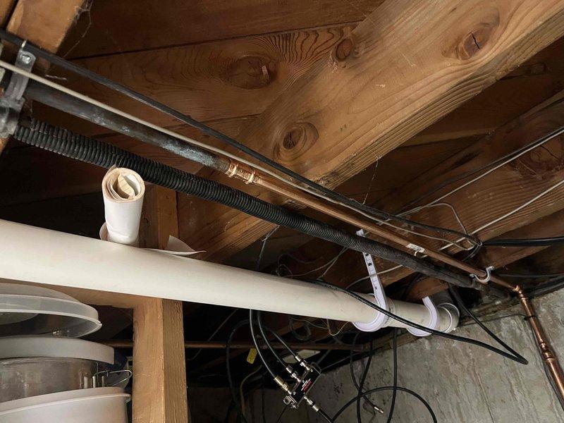 Completed plumbing project installation in basement utility area involving copper pipe installation with proper mounting brackets secured to concrete ceiling. Installed white PVC drainage system with vertical and horizontal runs, properly supported with straps and clips attached to wooden framing members. Coordinated piping routing alongside existing electrical conduit, black flexible tubing, and garage door track system to ensure proper clearances and accessibility for future maintenance.