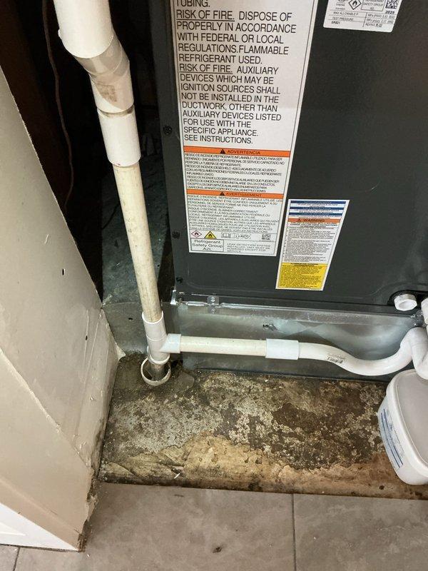 Responded to water backup at furnace location involving less than one-year-old EnergiAir system with warranty through May 2035. Upon inspection, found significant water accumulation and moisture damage on concrete floor surrounding HVAC unit and condensate drain assembly. Observed deteriorated floor conditions with visible water staining, debris accumulation, and organic growth patterns indicating ongoing drainage issue. Examined white PVC condensate drain piping configuration running vertically along unit and horizontally across floor, noting discoloration and dirt buildup on pipe surfaces. Documented floor flange connection point at base of vertical drain pipe showing dark discoloration and potential blockage indicators. Photographed extent of water damage, affected flooring, and compromised drain system components. System's current heating and cooling operational status could not be confirmed during visit due to water backup condition requiring immediate attention to drainage infras