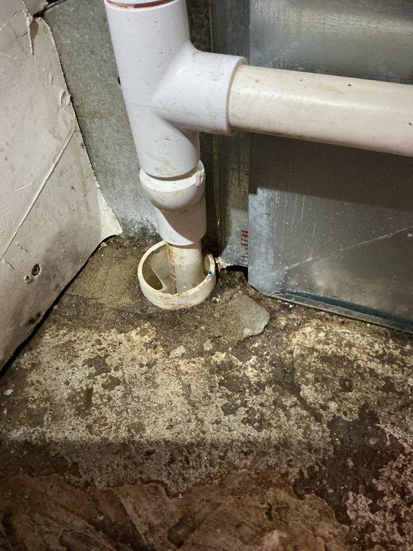 Responded to water backup at furnace location involving less than one-year-old EnergiAir system with warranty through May 2035. Upon inspection, found significant water accumulation and moisture damage on concrete floor surrounding HVAC unit and condensate drain assembly. Observed deteriorated floor conditions with visible water staining, debris accumulation, and organic growth patterns indicating ongoing drainage issue. Examined white PVC condensate drain piping configuration running vertically along unit and horizontally across floor, noting discoloration and dirt buildup on pipe surfaces. Documented floor flange connection point at base of vertical drain pipe showing dark discoloration and potential blockage indicators. Photographed extent of water damage, affected flooring, and compromised drain system components. System's current heating and cooling operational status could not be confirmed during visit due to water backup condition requiring immediate attention to drainage infras