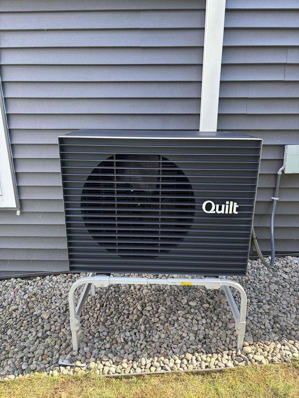 Completed installation of ductless mini-split system including outdoor Quilt brand condenser unit mounted on elevated stand with gravel pad and indoor wall-mounted air handler positioned above window. Installed refrigerant line set, electrical connections, and condensate drainage with all components tested and operational per manufacturer specifications.