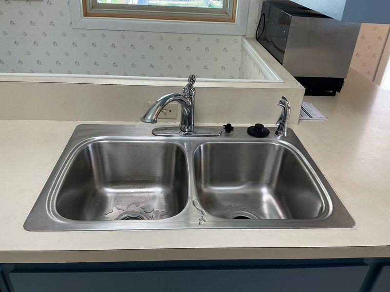 Completed installation of stainless steel double-basin kitchen sink with single-handle faucet, soap dispenser, and spray nozzle attachment. Under-sink plumbing configured with PVC drainage system including P-trap, dual chrome water supply lines with shut-off valves, and garbage disposal unit with electrical connection properly installed and tested for functionality.
