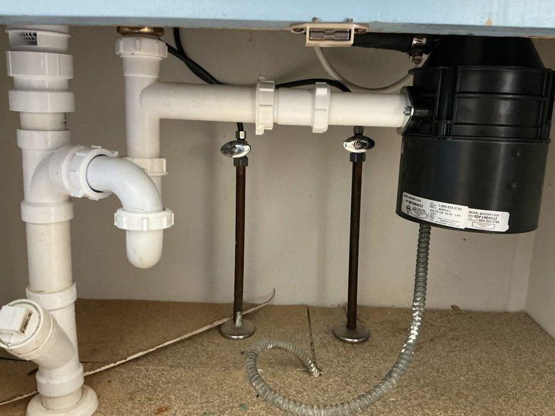 Completed installation of stainless steel double-basin kitchen sink with single-handle faucet, soap dispenser, and spray nozzle attachment. Under-sink plumbing configured with PVC drainage system including P-trap, dual chrome water supply lines with shut-off valves, and garbage disposal unit with electrical connection properly installed and tested for functionality.