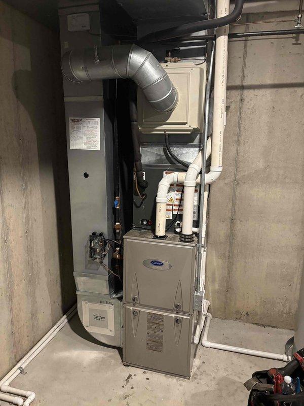 Completed full air conditioning system replacement including outdoor condensing unit and indoor air handler. Installed new Carrier equipment with proper refrigerant line connections, electrical conduit routing through exterior siding, and secure mounting on concrete pad. Unit positioned in landscaped side yard area with decorative rock placement maintained around base. Indoor installation included air handler integration with existing furnace system in basement utility area, new metallic ductwork with curved elbow transition, and proper sealing of all connections. Verified all electrical connections, tested system operation, and confirmed proper airflow throughout. Installation completed as same-day service per authorized work order.