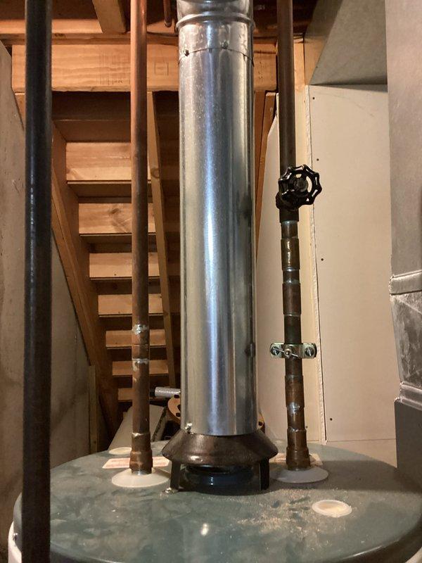 Conducted on-site evaluation of existing 40-gallon conventional tank water heater for replacement estimate in 27-year-old residential property. Inspected current water heater installation in basement utility area, documenting existing venting system with metal ductwork extending through wooden floor joists, copper supply line connections with shutoff valve, and concrete base platform. Photographed installation configuration including vertical venting arrangement and plumbing connections to provide accurate replacement specifications for standard tank-type water heater unit.