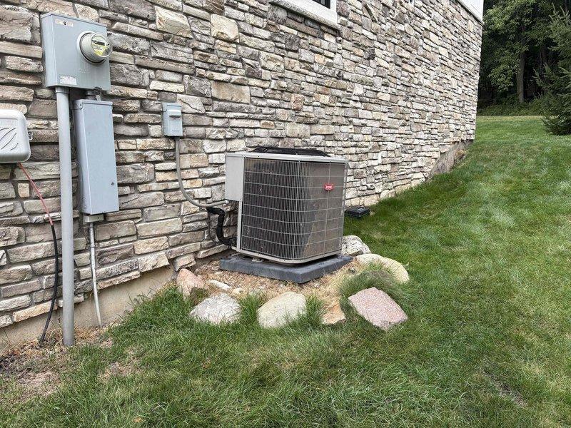 Responded to client request for assessment of unlevel air conditioning condenser unit. Upon arrival, performed visual inspection of 9-year-old Bryant residential AC system installed on concrete pad against exterior stone wall. Documented current installation conditions showing condenser unit settling unevenly on pad, with unit positioned on black rubber isolation pad surrounded by decorative river rock landscaping. Verified electrical disconnect box and conduit connections properly secured to wall. Photographed equipment positioning, pad condition, and surrounding grade to document extent of settling and provide basis for leveling estimate. Advised client that uneven condenser placement can lead to compressor damage, reduced efficiency, and premature system failure if not corrected. Provided detailed estimate for leveling service to restore proper equipment installation and prevent future mechanical issues.
