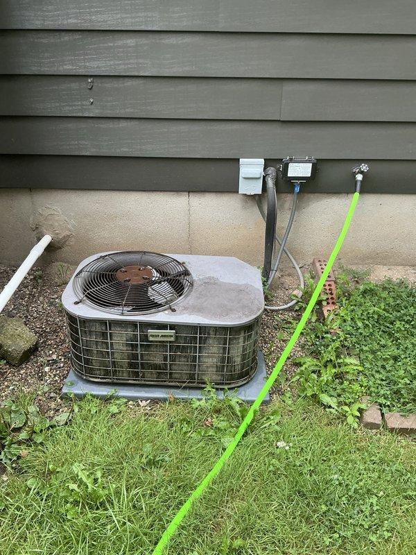 Conducted on-site evaluation of non-operational residential air conditioning system for complete unit replacement. Assessed existing 16-year-old outdoor condenser unit with beige metal casing and circular fan grille, positioned on ground-level pad against building exterior with electrical disconnect box and conduit connections. Documented interior electrical access panel and ceiling penetration points for system wiring routing. Provided comprehensive replacement estimate for new HVAC system installation.