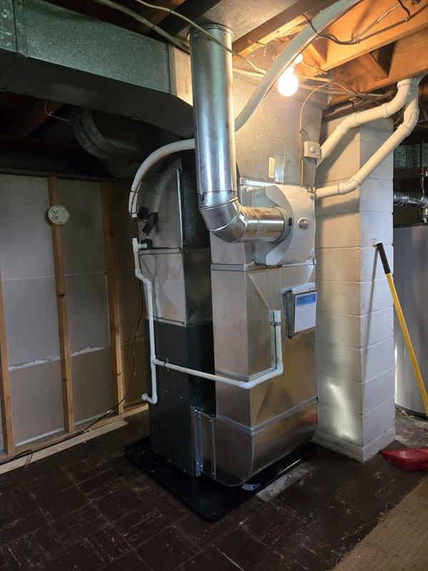 Completed full furnace installation in basement mechanical room. Installed modern gas heating system with dark cabinet unit, connected metal supply and return ductwork to existing distribution network, and routed metallic flue pipe vertically through ceiling for proper venting. Installed white PVC piping for condensate drainage and combustion air intake, ran electrical conduit and wiring to unit, and verified all connections met code requirements. System was tested for proper operation, airflow verified through duct network, and installation area left clean and organized.