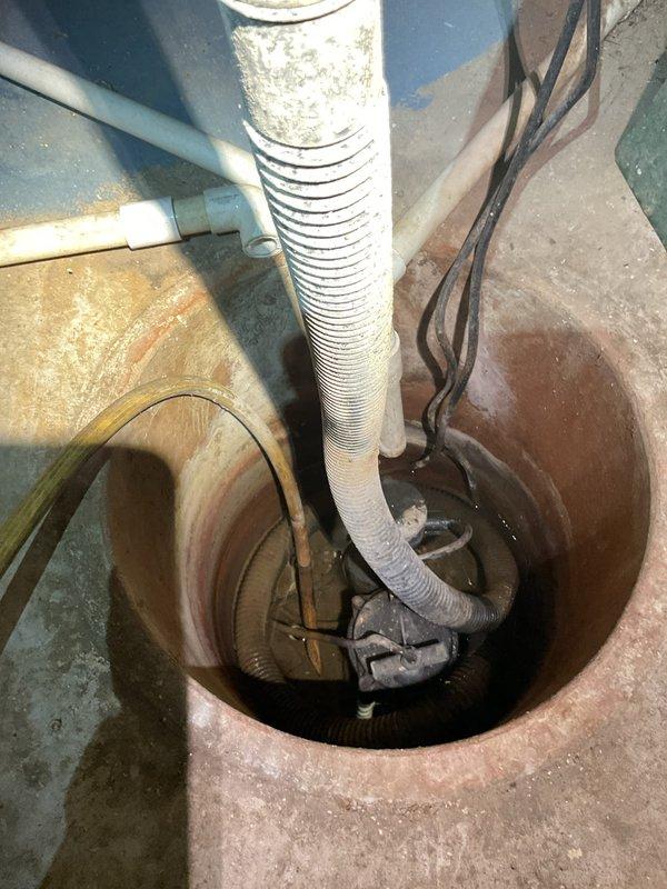 Responded to complaint of sump pump making intermittent noise in basement near water heater for past 1-2 weeks. Inspected sump pump assembly in concrete floor basin and found system operational but showing significant age-related wear consistent with forty-year-old home. Basin exhibited heavy rust staining and mineral deposits on walls indicating prolonged water exposure. Pump components, discharge piping, and electrical connections displayed visible discoloration and signs of moisture exposure over time. Documented existing conditions with photos showing white corrugated discharge hose, electrical wiring secured with cable ties, and overall pump assembly configuration. System currently functioning for water removal but displaying typical wear patterns that may warrant future replacement consideration.