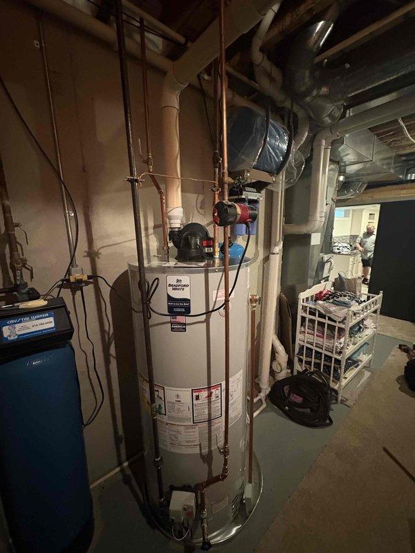 Completed installation of new residential water heater in basement utility area. Installed white cylindrical water heater with recirculation pump mounted on top, connected all copper supply and distribution piping with isolation valves, and integrated unit with existing blue expansion tank and dual-stage water filtration system. All connections tested and verified for proper operation with no leaks detected.