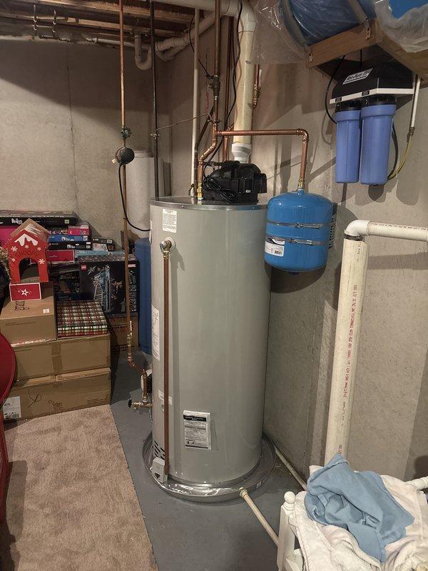 Completed installation of new residential water heater in basement utility area. Installed white cylindrical water heater with recirculation pump mounted on top, connected all copper supply and distribution piping with isolation valves, and integrated unit with existing blue expansion tank and dual-stage water filtration system. All connections tested and verified for proper operation with no leaks detected.