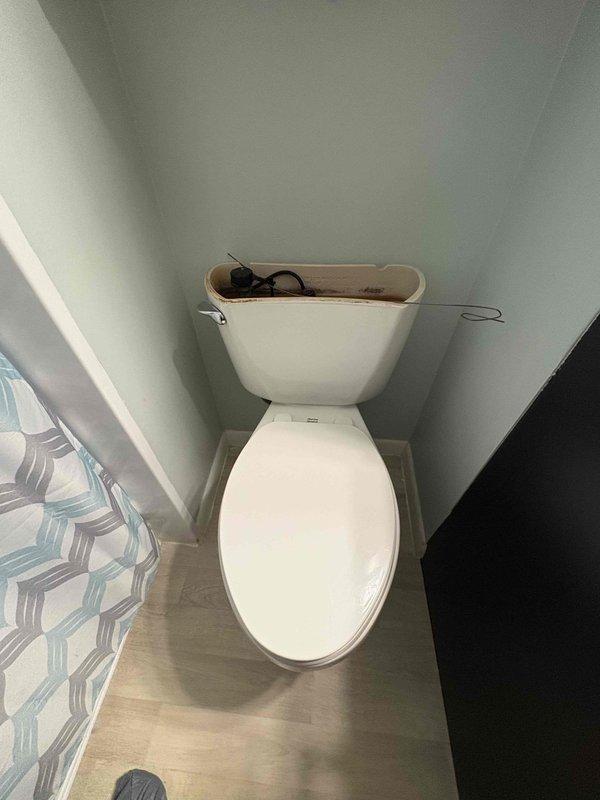 Responded to service call for toilet replacement in 1958-built home. Upon arrival, inspected non-functional toilet that had been out of service for several weeks. Documented existing conditions showing white porcelain toilet with exposed tank mechanism and black decorative water supply line in light blue-green painted bathroom with laminate flooring. Photographed plumbing connections including shut-off valve and supply line configuration for records. Toilet remained out of service with alternative bathroom facilities available to occupants.