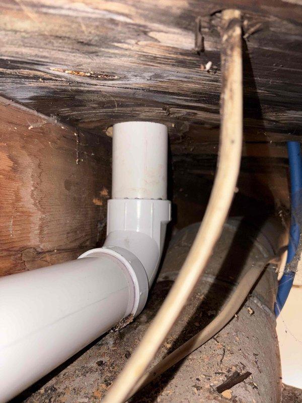 Completed kitchen drain rebuild involving replacement of deteriorated PVC drain piping in crawlspace area beneath kitchen. Installed new white PVC drain line with 90-degree elbow fittings to restore proper drainage flow, working around existing copper water supply lines and aged floor joists. Vertical drain stack connection completed where pipe transitions through floor structure to tie into existing plumbing system.