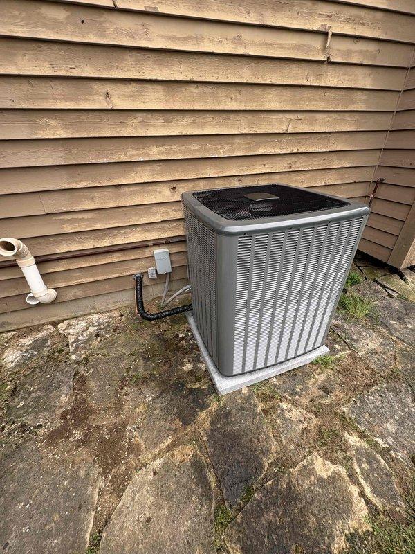 Complete residential air conditioning system replacement performed. Removed existing outdoor condensing unit and indoor air handler equipment. Installed new outdoor AC condenser on existing concrete pad with proper leveling and positioning against building exterior. Secured refrigerant line sets and electrical conduit from exterior wall to new condenser unit. Utilized existing PVC condensate drain lines for proper drainage. Indoor installation included new air handler unit positioned in basement utility area with integration to existing ductwork system. Connected all electrical components, refrigerant lines, and condensate drainage. Verified proper thermostat wiring and digital display functionality. Pressure tested refrigerant lines for leaks, evacuated system, and charged to manufacturer specifications. Performed system startup procedures and verified proper operation of both heating and cooling functions. Confirmed adequate airflow through supply ductwork and proper temperature diff