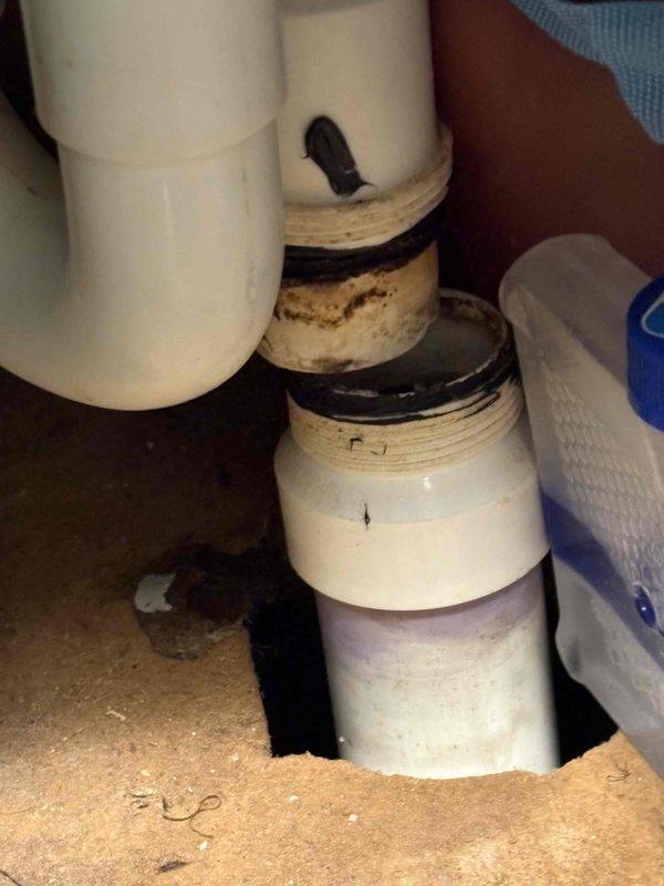 Responded to report of slow drip in basement originating from main floor plumbing, ongoing for approximately six months. Inspected under-sink plumbing assembly and identified source of leak at threaded PVC drain connection with deteriorated rubber gasket. Found significant age-related wear on drain pipes with discoloration and grime buildup around joints and connection points, consistent with original 1950 home construction. Documented condition of plumbing fixtures with photographs showing compromised seals and moisture damage to surrounding cabinet surfaces. Evaluated water softener as requested and found unit operational. Provided recommendations for replacement of worn gaskets and drain assembly components to resolve active leak and prevent future water damage.