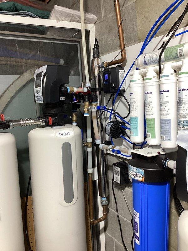 Conducted comprehensive plumbing project estimate for reverse osmosis water filtration system installation in utility room. Assessed existing multi-stage water treatment infrastructure including NUVO water softener, reverse osmosis filtration assembly with multiple white cylindrical filter cartridges, blue pressure tank, and complex plumbing network featuring copper pipes, PVC connections, brass valves, and electronic control devices with LED indicators. Documented current system configuration and prepared estimate for customer water quality optimization project.