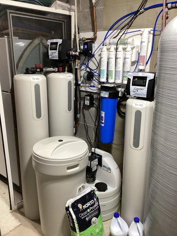 Conducted comprehensive plumbing project estimate for reverse osmosis water filtration system installation in utility room. Assessed existing multi-stage water treatment infrastructure including NUVO water softener, reverse osmosis filtration assembly with multiple white cylindrical filter cartridges, blue pressure tank, and complex plumbing network featuring copper pipes, PVC connections, brass valves, and electronic control devices with LED indicators. Documented current system configuration and prepared estimate for customer water quality optimization project.