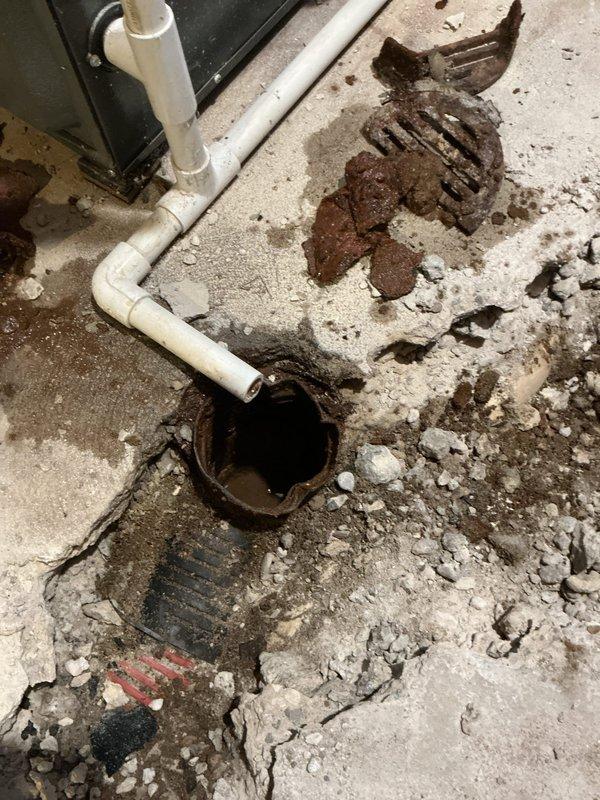 Replaced deteriorated cast iron drain line located beneath concrete slab in basement utility area. Removed failed section of cast iron piping that showed significant corrosion and breakdown, then installed new PVC drainage system to restore proper function. Concrete floor exhibited extensive moisture damage, cracking, and deterioration around the drain opening, with visible standing water and rust staining indicating long-term drainage issues that necessitated the underground pipe replacement.