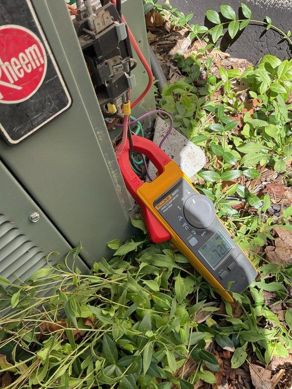 Responded to no cooling complaint on 20+ year old Rheem outdoor condensing unit. Upon arrival, found unit running but not cooling to set temperature. Performed electrical diagnostics using clamp meter at outdoor unit's electrical connection points. Tested voltage and amperage readings at compressor and condenser fan motor circuits. Inspected wire connections including red, orange, green, purple, and yellow conductors entering unit through electrical enclosure. Unit showed significant age-related weathering, dirt accumulation, and vegetation overgrowth around base affecting airflow. Documented electrical measurements and overall equipment condition with photos. Unit's advanced age (20+ years) indicates likely refrigerant loss, compressor degradation, or failed capacitor requiring further diagnosis and repair recommendations.