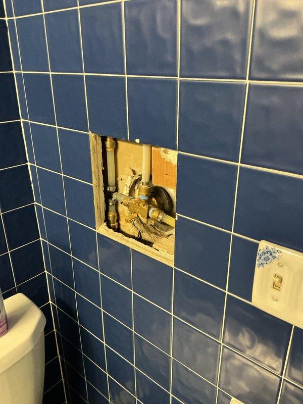 Responded to a plumbing leak originating from the shower area and penetrating through to the basement below. Created an access panel in the tiled bathroom wall to expose the concealed plumbing system. Upon inspection, identified the source of the leak at the shower valve assembly behind the navy blue tiled enclosure. The copper supply lines and brass valve body showed signs of moisture intrusion and aging that was causing water to bypass the fixture and travel downward through the floor structure. Assessed the condition of the valve assembly, surrounding framing, and water damage extent. Documented all findings with photographic evidence of the tiled shower interior, valve location, and exposed plumbing components for repair recommendations.