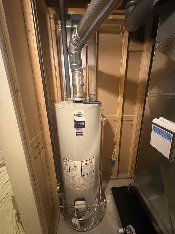 Completed full installation of new HVAC system and water heater in residential utility space. Installed high-efficiency furnace with diamond-pattern exterior casing, mounted on protective drip pan with integrated thermostat control panel and blue display screen. Ran white PVC venting from furnace unit vertically through ceiling, installed electrical conduit and necessary piping connections along unit perimeter. Installed white cylindrical water heater with aluminum flex duct venting routed upward to ceiling exhaust point, connected all required plumbing lines and safety components. Both units positioned in unfinished basement area with exposed wall framing, all installations completed per manufacturer specifications and local code requirements. System tested and verified operational upon completion.