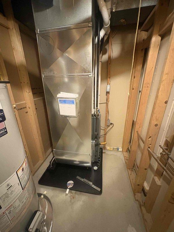 Completed full installation of new HVAC system and water heater in residential utility space. Installed high-efficiency furnace with diamond-pattern exterior casing, mounted on protective drip pan with integrated thermostat control panel and blue display screen. Ran white PVC venting from furnace unit vertically through ceiling, installed electrical conduit and necessary piping connections along unit perimeter. Installed white cylindrical water heater with aluminum flex duct venting routed upward to ceiling exhaust point, connected all required plumbing lines and safety components. Both units positioned in unfinished basement area with exposed wall framing, all installations completed per manufacturer specifications and local code requirements. System tested and verified operational upon completion.