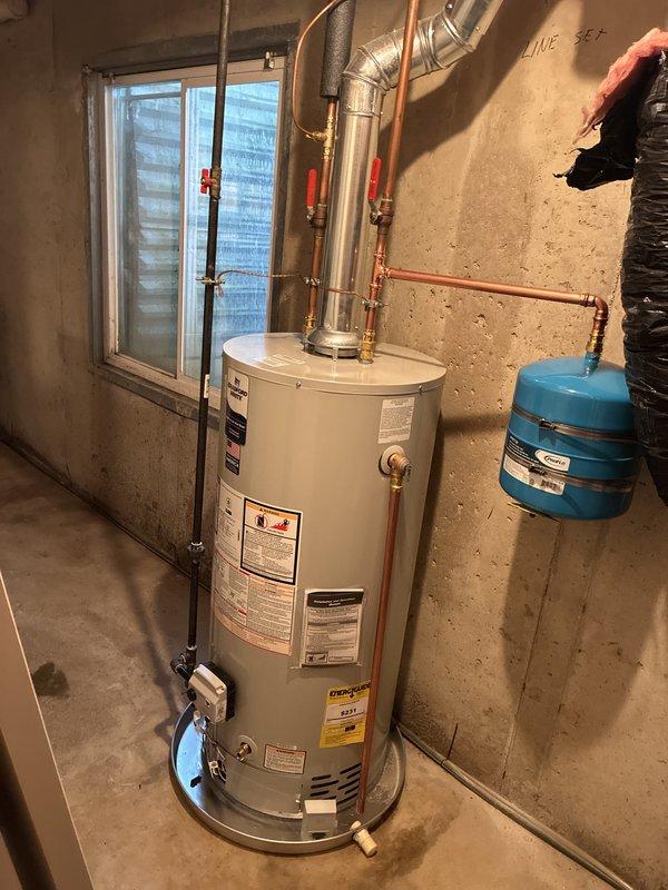 Completed full water heater installation in basement utility room. Removed existing unit and installed new tall cylindrical water heater with proper venting connections to existing ductwork system. Connected copper supply lines with red-handled isolation valves for system control and installed blue expansion tank on wall for thermal expansion protection. Verified all plumbing connections, tested pressure relief valve operation, and ensured proper clearances from furnace and other mechanical equipment. System tested and operational.