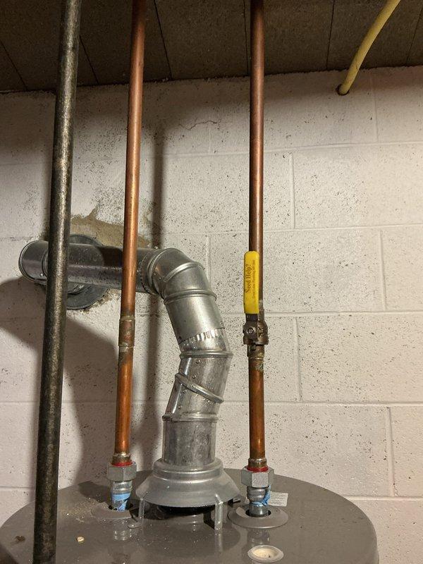 Conducted estimate for water heater replacement in residential basement due to active leak from existing unit. Inspected current gray cylindrical water heater with galvanized and copper supply piping, exhaust venting through ceiling, and yellow-handled shut-off valve. Documented significant rust-colored staining and water damage on concrete block wall around pipe penetration, indicating chronic leakage. Photographed existing plumbing configuration including three vertical pipes, metallic elbow joint connection to flue pipe, and deteriorated pipe penetration point with visible mineral deposits and corrosion on metal components. Provided replacement options and recommendations for new water heater installation.
