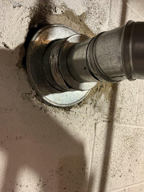 Conducted estimate for water heater replacement in residential basement due to active leak from existing unit. Inspected current gray cylindrical water heater with galvanized and copper supply piping, exhaust venting through ceiling, and yellow-handled shut-off valve. Documented significant rust-colored staining and water damage on concrete block wall around pipe penetration, indicating chronic leakage. Photographed existing plumbing configuration including three vertical pipes, metallic elbow joint connection to flue pipe, and deteriorated pipe penetration point with visible mineral deposits and corrosion on metal components. Provided replacement options and recommendations for new water heater installation.