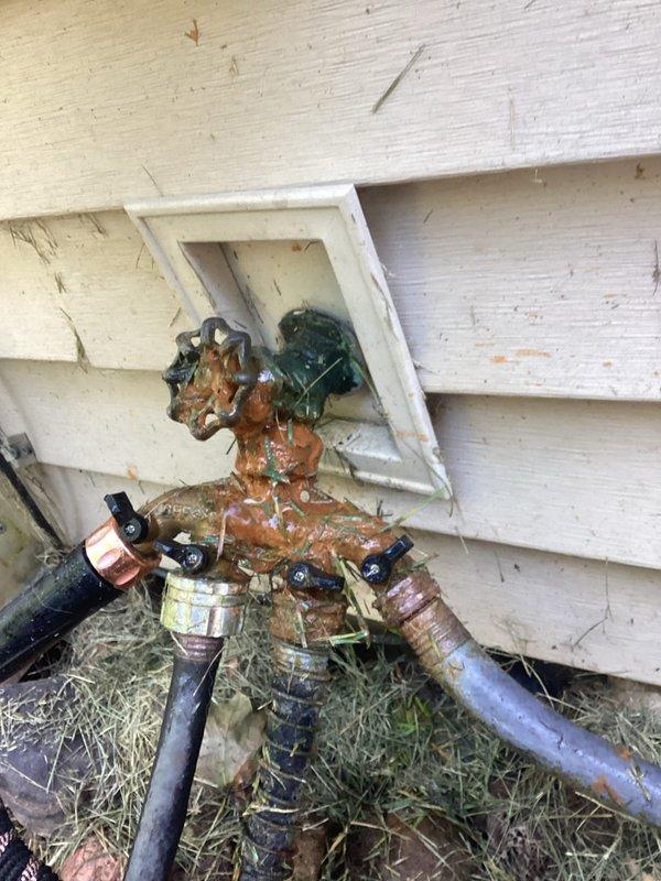 Responded to service call for intermittent outdoor faucet leak at forty-year-old residence. Upon inspection, discovered exterior hose bib and valve assembly with extensive rust and corrosion throughout all metal components, including valve body, Y-connector splitter, and fittings. Heavy orange-brown oxidation and white mineral deposits indicated prolonged water exposure and lack of maintenance. Valve handle showed green patina consistent with aged brass or bronze material. Black rubber hoses connected to corroded assembly showed signs of weathering. Debris accumulation at base of unit and staining on adjacent siding confirmed active leak conditions. Documented condition with photographs of exterior valve assembly and interior plumbing connections visible in utility area. Interior inspection revealed copper supply lines and mounting configuration against pegboard wall system. Recommended complete replacement of outdoor faucet assembly due to severity of corrosion and compromised integri