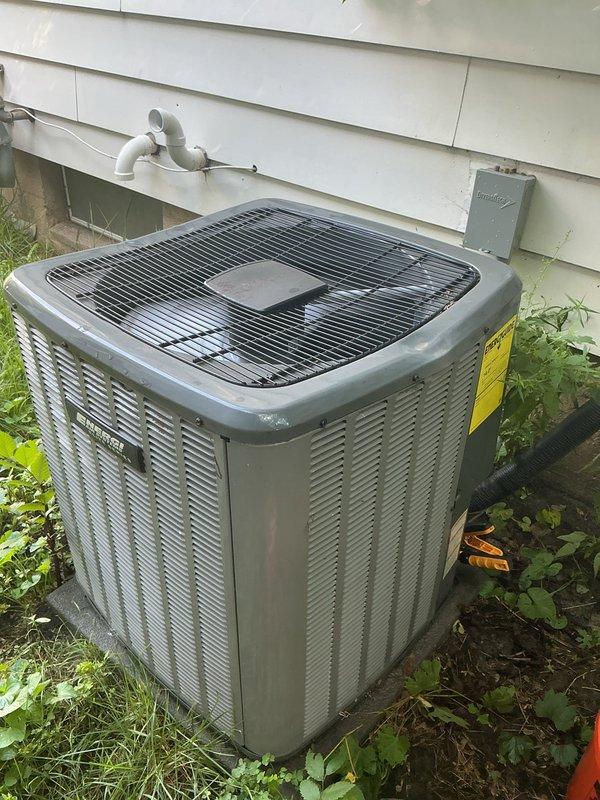 Completed annual cooling system maintenance and inspection on residential HVAC unit. Accessed equipment through rear alley entrance, inspected outdoor condenser unit with proper clearances on grass pad adjacent to building with white vinyl siding, verified electrical disconnect box operation, and examined refrigerant line insulation and PVC drainage routing. Indoor evaporator coil and refrigerant lines inspected behind appliance installation showing proper insulation on copper lines and secure connections. All components found operating within normal parameters, system tuned and ready for cooling season.