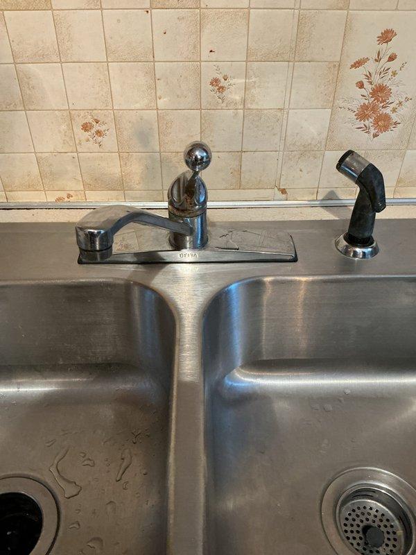Responded to kitchen faucet repair call at a 40-year-old residential property where issues had persisted for three days. Inspected chrome single-handle ball-style faucet assembly with separate spray nozzle on stainless steel double-basin sink. Diagnosed and repaired faulty faucet components, restoring proper water flow and eliminating leaks. Documented existing conditions including aged tile backsplash with visible staining and discoloration around fixture area.