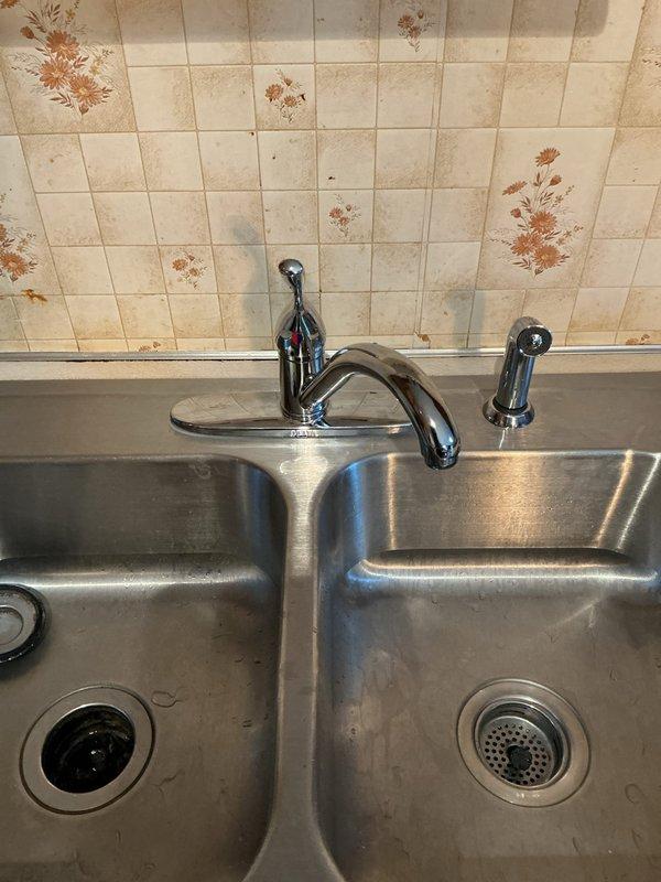 Responded to kitchen faucet repair call at a 40-year-old residential property where issues had persisted for three days. Inspected chrome single-handle ball-style faucet assembly with separate spray nozzle on stainless steel double-basin sink. Diagnosed and repaired faulty faucet components, restoring proper water flow and eliminating leaks. Documented existing conditions including aged tile backsplash with visible staining and discoloration around fixture area.