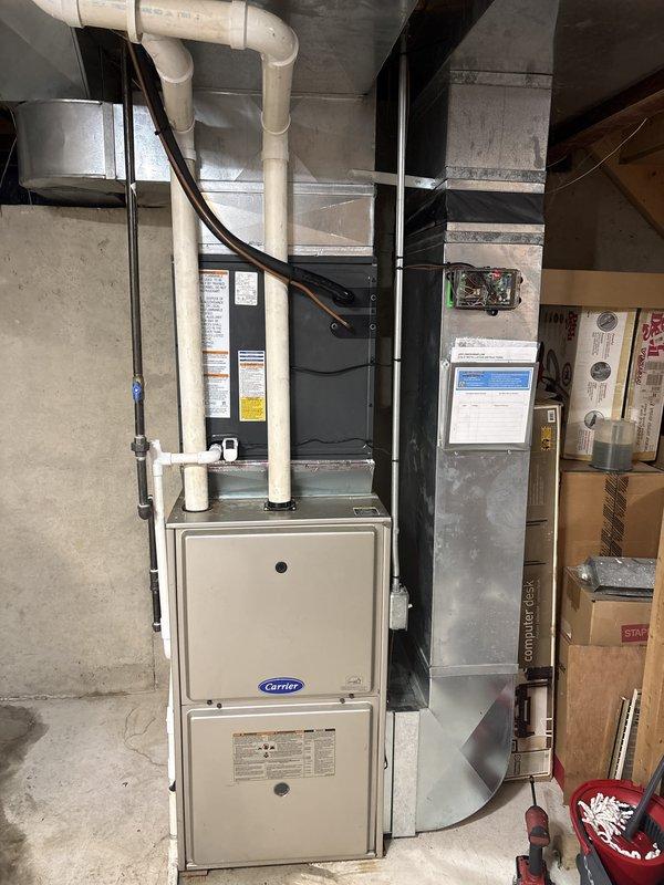 Successfully completed installation of new Carrier HVAC system and residential water heater in basement utility area. The HVAC installation included proper mounting of furnace unit with air handler, installation of PVC venting pipes, and electrical conduit connection. The water heater replacement involved installing a new white cylindrical unit with copper supply piping, brass fittings, and flexible conduit for electrical connection. Both units were positioned against concrete walls on concrete flooring, with all connections properly secured and tested for operation.