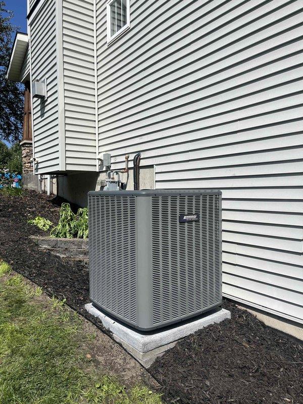 Completed full air conditioning system replacement at residential property. Removed existing outdoor condensing unit and installed new gray-green AC condenser on concrete pad against exterior wall with white vinyl siding. Ran refrigerant lines with black insulation from unit up exterior wall. Installed electrical disconnect box on concrete mounting pad with proper conduit connections to condensing unit. Verified all electrical connections, tested system operation, and confirmed proper refrigerant charge levels. Landscaped area around installation with fresh mulch and restored ground cover.