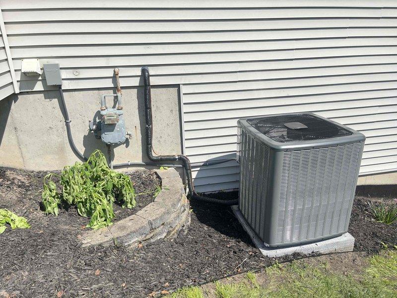 Completed full air conditioning system replacement at residential property. Removed existing outdoor condensing unit and installed new gray-green AC condenser on concrete pad against exterior wall with white vinyl siding. Ran refrigerant lines with black insulation from unit up exterior wall. Installed electrical disconnect box on concrete mounting pad with proper conduit connections to condensing unit. Verified all electrical connections, tested system operation, and confirmed proper refrigerant charge levels. Landscaped area around installation with fresh mulch and restored ground cover.