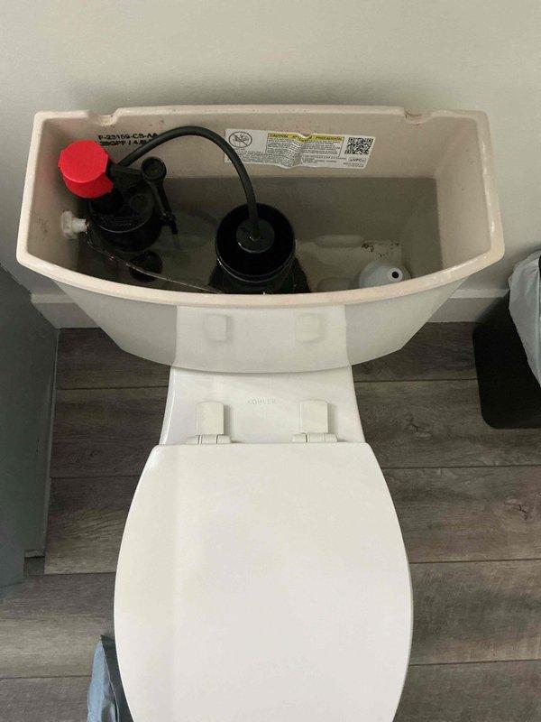 Responded to customer complaint of vibrating noise occurring during toilet flush cycle in 32-year-old home. Upon inspection, removed tank lid and examined internal flush valve assembly, fill valve with red cap, flapper mechanism, and refill tube connections. Identified worn fill valve as likely source of vibration noise. Photographed tank components showing black plastic flush valve assembly, red-capped fill valve on left side, rubber flapper with chain attachment, and connecting refill tube for documentation purposes. No active leaks observed at time of service call.