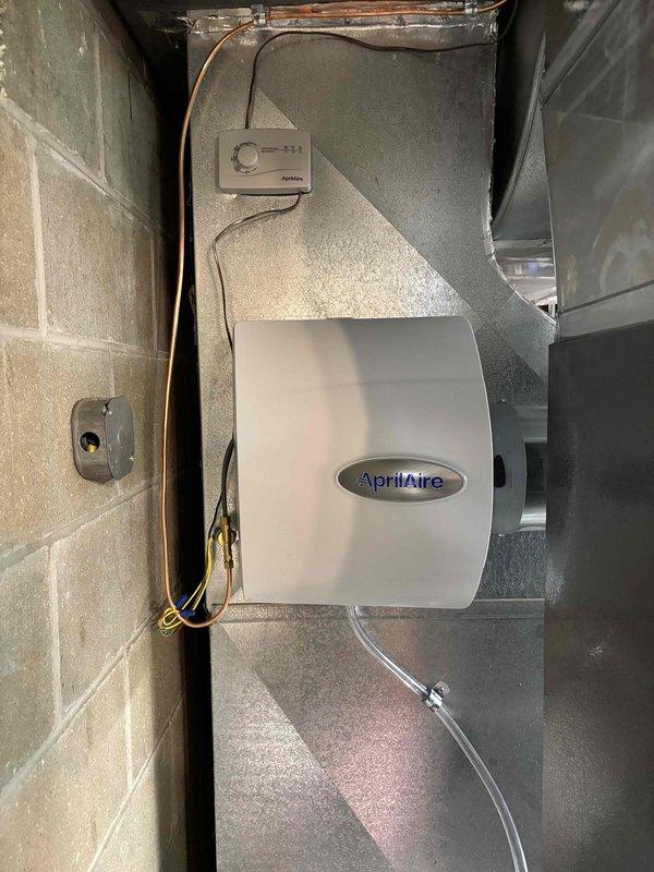Completed full HVAC system installation including new furnace and air conditioning condenser unit. Installed outdoor AC condenser on composite pad adjacent to home foundation with proper refrigerant line connections and electrical disconnect box mounted to structure. Installed AprilAire whole-home humidifier unit integrated with ductwork system in basement utility area, including humidistat control panel, water supply line with copper tubing, drainage connections, and electrical wiring. All components properly mounted, connected, and positioned for optimal system performance and accessibility for future maintenance.