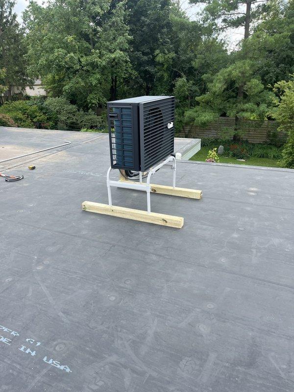 Completed full installation of ductless mini-split system at residential property. Outdoor condenser unit was mounted on reinforced white metal stand with wooden support beams on flat rubber membrane rooftop, positioned near corner of building adjacent to existing gutter and downspout system. Indoor and outdoor units were successfully installed, connected, and tested for proper operation.