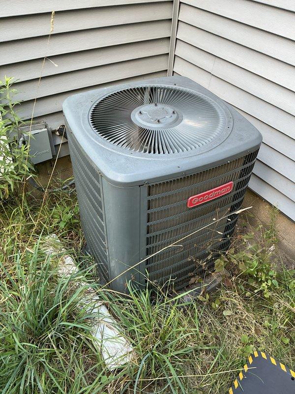 Responded to cooling complaint on 8-year-old Goodman system where air was flowing from vents but house was not cooling adequately. Upon arrival, found outdoor condenser unit surrounded by overgrown vegetation and debris restricting airflow through side louvers. Customer had already replaced filter and hosed down unit prior to service call. Performed visual inspection of system components, confirmed no electrical trips, verified normal airflow at registers, and observed no frost on refrigerant lines. Cleared vegetation and debris from around condenser perimeter to restore proper airflow clearance. Documented equipment conditions with photos showing vegetation encroachment and overall system setup for service records.